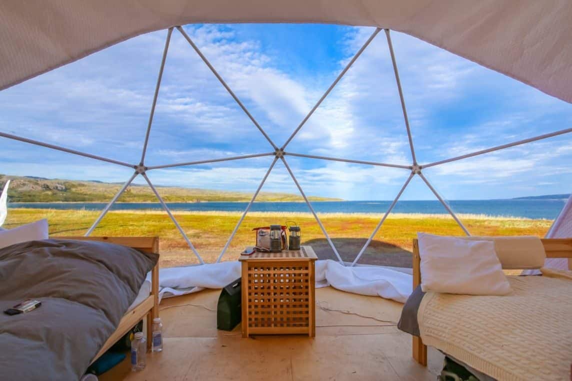 view of a lake from the inside of a dome shaped cabin with 2 beds and end table between them