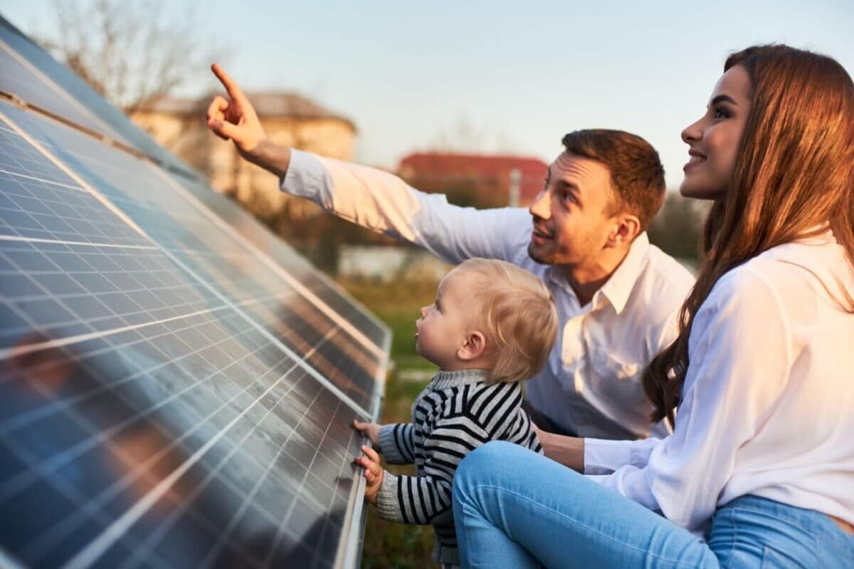 Family consisting of man, woman, and small child sitting and smiling in front of solar panel, man pointing to something in the distance with everyone looking in that direction.
