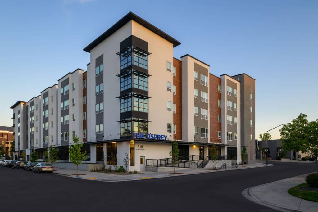 Modern four-story student housing building with podium parking, large windows, and a mix of white, gray, and wood-toned exterior finishes, located at 818 Ann Morrison Park Drive in Boise, Idaho