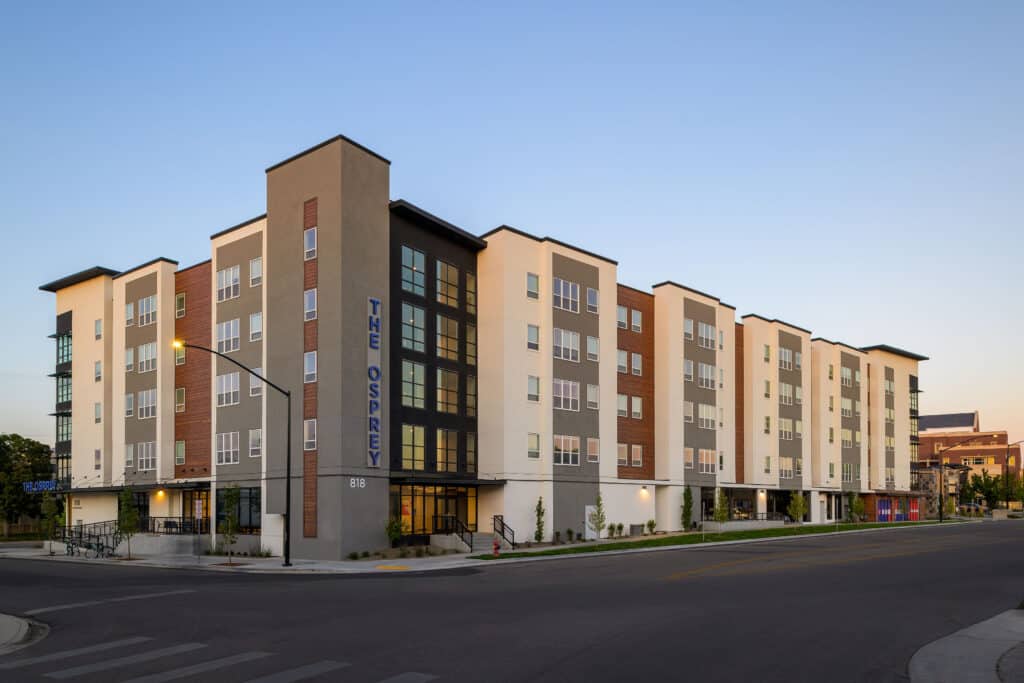 Modern four-story student housing building with podium parking, large windows, and a mix of white, gray, and wood-toned exterior finishes, located at 818 Ann Morrison Park Drive in Boise, Idaho