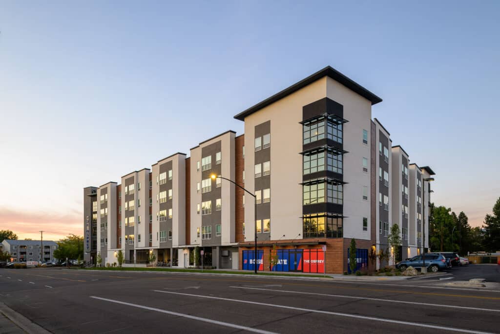 Modern four-story student housing building with podium parking, large windows, and a mix of white, gray, and wood-toned exterior finishes, located at 818 Ann Morrison Park Drive in Boise, Idaho