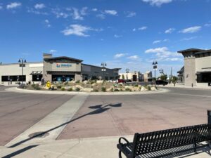 Outdoor view of a modern shopping center with a roundabout in front, featuring the Sparkzone Athletics building and other retail stores under a blue sky with scattered clouds.